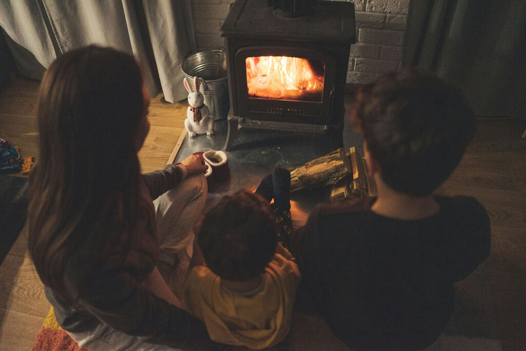 A woman and two children sit in front of a lit wood stove.