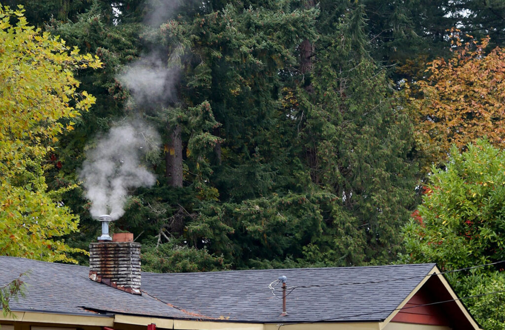 A roofline view of a house’s chimney emitting smoke, with trees in the background.